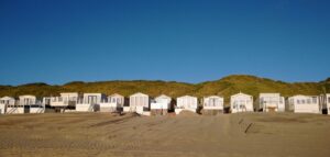 Tekst alternatywny: Domki plażowe ustawione w rzędzie na piaszczystej plaży, przed zielonymi wydmami, przypominające o tym, jak inna niż budowa domów stalowych może być architektura wypoczynkowa.