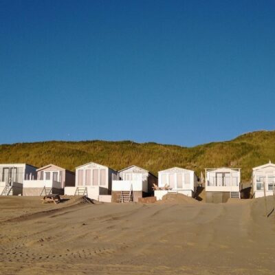 Tekst alternatywny: Domki plażowe ustawione w rzędzie na piaszczystej plaży, przed zielonymi wydmami, przypominające o tym, jak inna niż budowa domów stalowych może być architektura wypoczynkowa.