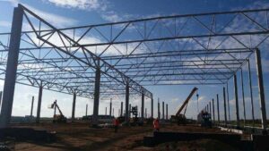 Konstrukcja stalowa budynku w trakcie montażu, gdzie zastosowanie technologii w budownictwie stalowym jest widoczne. Pracownicy przy użyciu dźwigu działają na placu budowy.