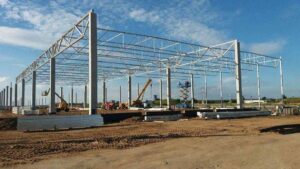 Na obrazku widoczny jest plac budowy dużej konstrukcji, prawdopodobnie hali stalowej.